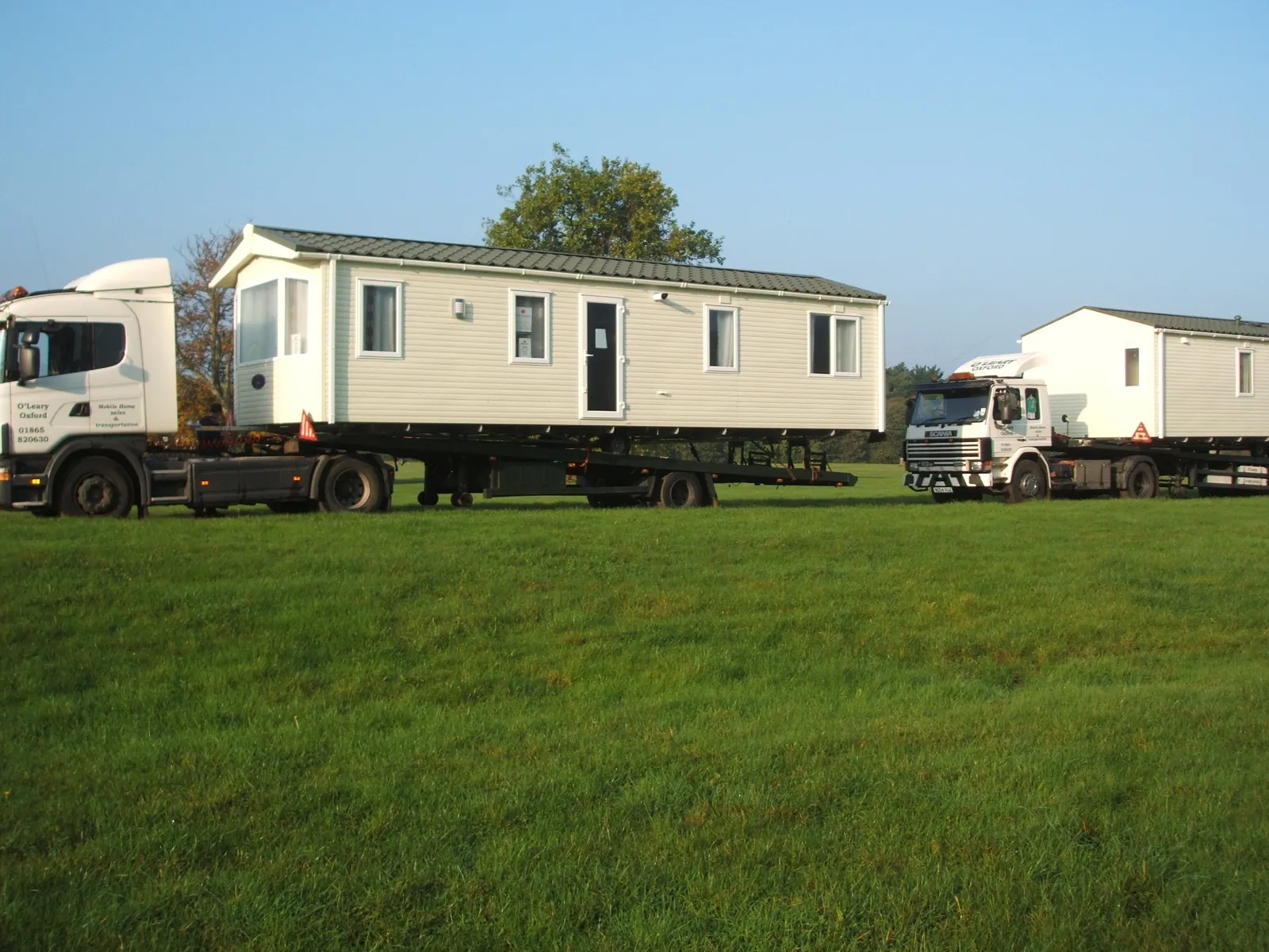 Two O'Leary lorries, each carrying a static caravan, on a grass field
